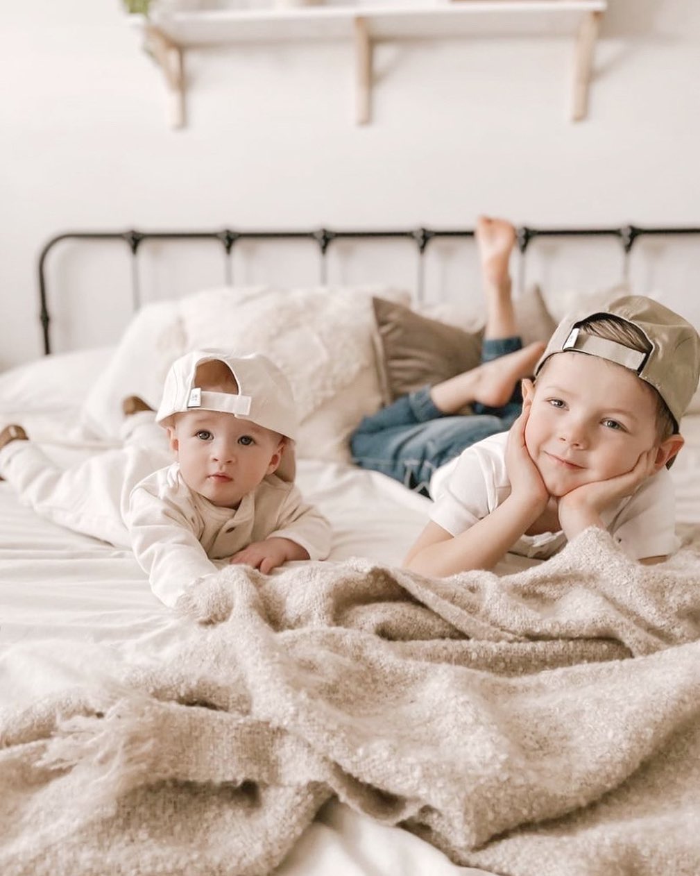 Two young brothers relaxing on the bed while wearing matching sun hats in different colours. Toddler is wearing a white hat, old child / kid is wearing a khaki green hat. Hats are a minimalist five-panel design, made in Canada out of organic cotton. Hats have an adjustable soft velcro closure. 