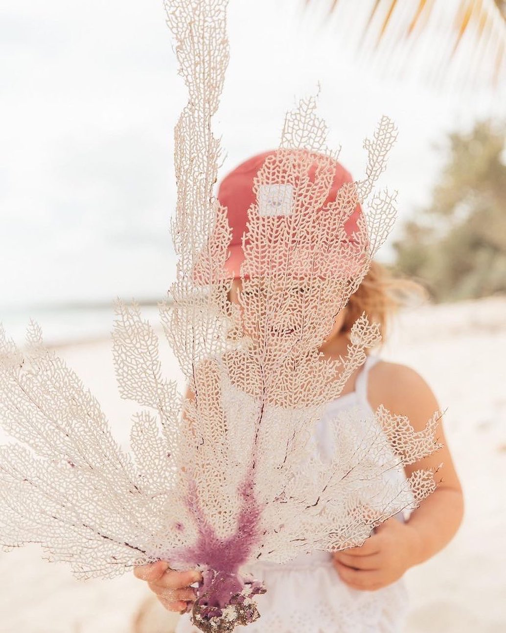 Toddler girl is on the beach playing with coral while wearing a pink / red sun hat. Hat is minimalist five-panel design, made in Canada out of organic cotton.